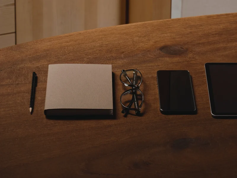 Illustrazione: occhiali da lettura, taccuino, matita, telefono e tablet su un tavolo di legno.